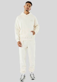 Giovane uomo che indossa una felpa crema e pantaloni della tuta abbinati, con scarpe da ginnastica bianche e nere, in piedi davanti a uno sfondo grigio uniforme.