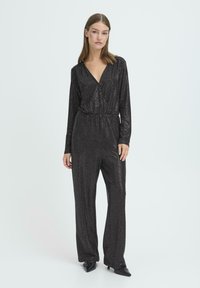 Czarny kombinezon z cekinami, z długimi rękawami i głębokim dekoltem w kształcie litery V. Posiada wyraźnie zaznaczoną talię i szerokie nogawki. Teksturowana, błyszcząca powierzchnia.