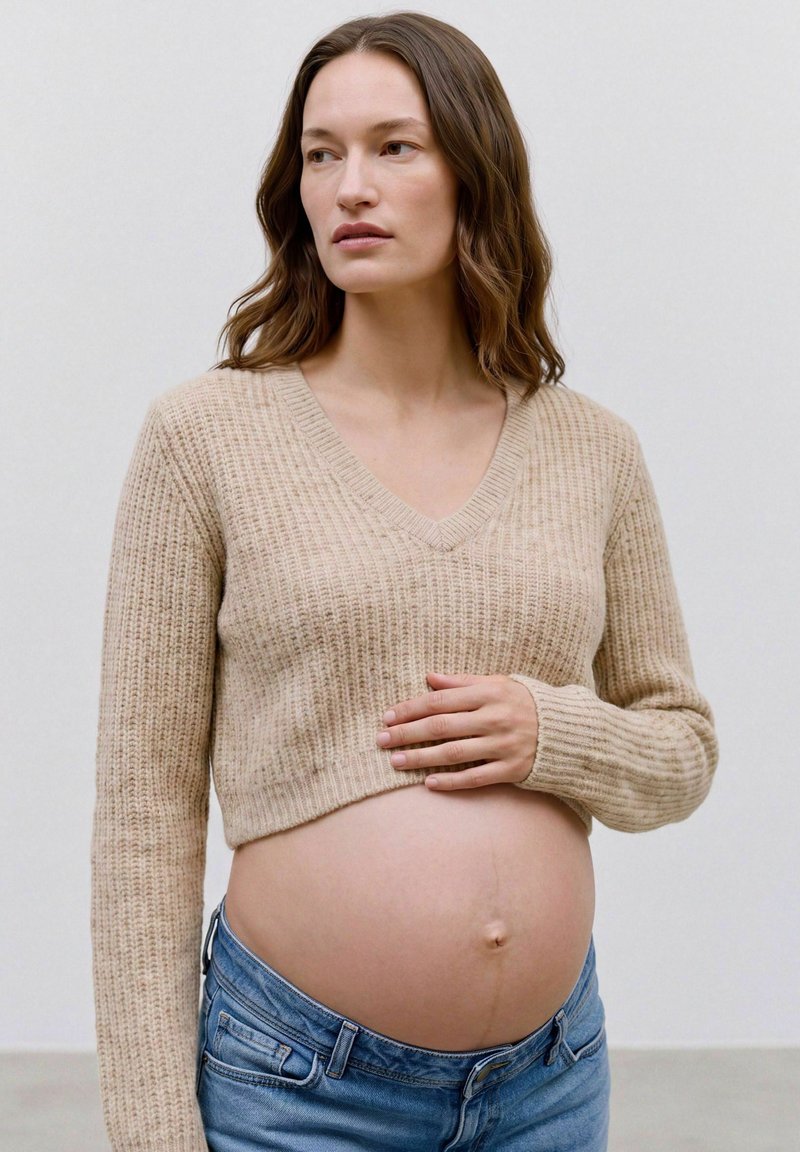 Femme enceinte aux longs cheveux bruns portant un pull court beige et un jean bleu, tenant son ventre nu et regardant sur le côté.