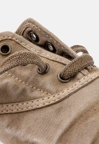 Chaussure en toile marron avec œillets en métal et lacets tissés. Présente une surface texturée et des détails de couture le long des bords.