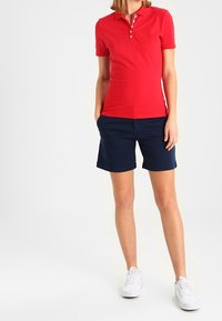 Červené polo tričko s límečkem a zapínáním na tři knoflíky, v kombinaci s námořnickými šortkami. Bílé tenisky dotvářejí outfit. Hladká textura látky.