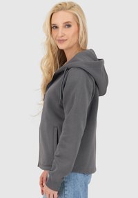 Felpa grigia con cappuccio, dalla texture liscia, con zip frontale, tasche laterali e polsini a coste. Indossata sopra un paio di jeans azzurri chiari.