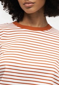 Γυναίκα με σγουρά μαλλιά που φορά λευκό και πορτοκαλί ριγέ πουκάμισο με πορτοκαλί γιακά, εμφανιζόμενη από τους ώμους μέχρι τα χείλη σε λευκό φόντο.