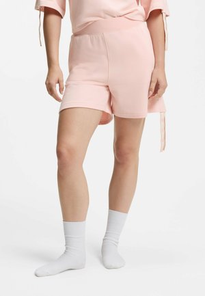 Persoon draagt lichtroze casual shorts, bijpassende top en witte sportsokken, staand tegen een effen lichte achtergrond.