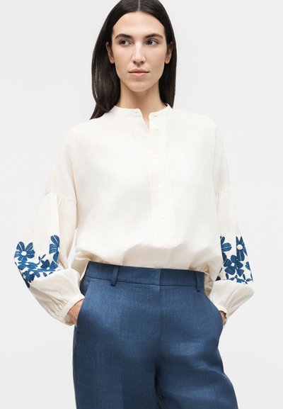 Γυναίκα που φοράει λευκό μπλουζάκι με μπλε κεντήματα με λουλούδια στα μανίκια και μπλε ψηλόμεσο παντελόνι, τα χέρια στις τσέπες, κοιτάζει πλάγια.