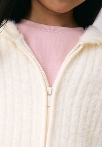 Veste zippée couleur crème en tissu texturé et duveteux avec des nervures verticales, portée par-dessus un haut rose clair à encolure ronde.
