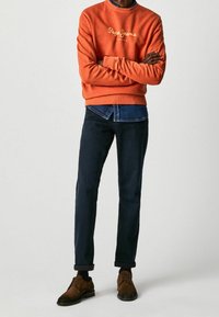 Oranžová mikina s logem "Pepe Jeans", tmavě modrá denimová košile pod ní, tmavě modré kalhoty a hnědé semišové boty.