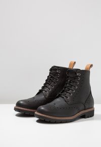 Stivaletti neri in pelle con dettagli brogue, punta tonda, chiusura con lacci e un accento sul tacco in tonalità marrone chiaro.