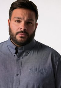 Camisa de botones azul oscuro hecha de un tejido texturizado, con un logotipo bordado de "SAILOR" que incluye coordenadas en el área del pecho izquierdo.