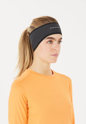 Svart hårband med prickig textur, med en logo, bärs av en person i en ljus orange långärmad topp.