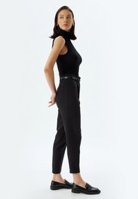 Zwarte ribbed mouwloze top gecombineerd met hoge taille zwarte broek en glanzende zwarte loafers. Kenmerken zijn onder andere een dunne riem en een getailleerde pasvorm.