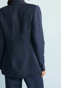 Blazer marine en tissu lisse, avec des épaules larges, une coupe ajustée et des accents de boutons sur les manches. Vue de dos montrée.