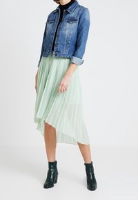 Žena oblečená v srolované džínové bundě, černém topu, světle zelené plisované asymetrické sukni a tmavě zelených kotníkových botách, stojící na bílém pozadí.