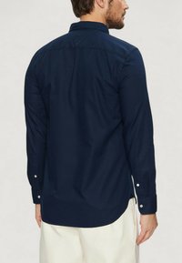 Maglietta a maniche lunghe navy in cotone, con colletto classico, polsini con bottoni e orlo arrotondato. La vista posteriore mostra una texture liscia e cuciture.