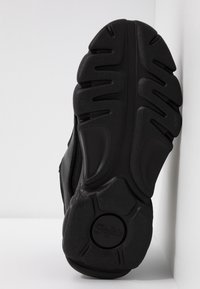 Černá podrážka tenisky s hlubokými drážkami a kruhovým vzorem běhounu, postavená vzpřímeně na bílém pozadí.