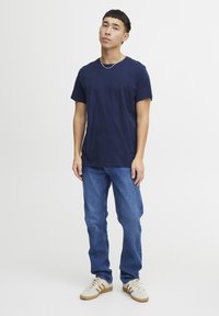 Modra bombažna majica s kratkimi rokavi in okroglim ovratnikom, v kombinaciji z modrimi kavbojkami in kremnimi športnimi copati z rjavimi akcenti.