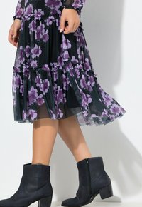 Frau in einem schwarzen und lila geblümten Midikleid und schwarzen Stiefeletten, die mit einer Hand den Saum des Kleides hält.