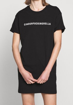 Osoba nosząca czarną sukienkę typu T-shirt z białym napisem "CINDERFUCKINGRELLA" na klatce piersiowej, dłonie złączone z przodu.