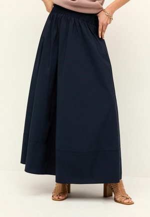 Femme portant une jupe bleu marine longueur cheville avec une ceinture élastique et des sandales beiges à talons ouvertes.