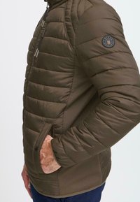 Mann iført olivengrønn quiltet jakke med glidelås på brystlommen, hånd i siden av trykknapp-lommen, og rundt logo på overarmen.