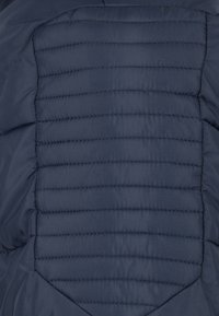 Sekce námořnicky modré péřové bundy s prošitým designem. Má vodorovné prošívání a hladkou texturu. Žádné viditelné kovové doplňky ani ozdoby.