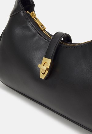 Sac à main en cuir noir avec fermeture éclair dorée et boucle tourniquet décorative dorée sur la sangle avant, incliné sur fond blanc.