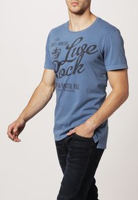 Modrá bavlněná trička s krátkým rukávem a kulatým výstřihem s černým potiskem "Live Rock." Nosené s tmavými džíny.