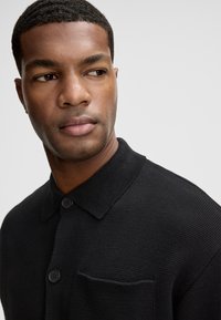 Czarna dzianinowa koszulka z kołnierzykiem, dwoma guzikami i kieszonką po lewej stronie. Tekstura wydaje się gładka i gęsta.