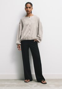 Bomber jacket grigio chiaro con ricami floreali, chiusura con zip e maniche a palloncino, abbinato a pantaloni neri a gamba larga e sandali.