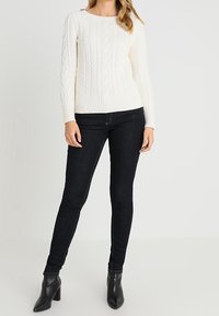 Pull blanc en maille torsadée avec ourlet côtelé, associé à un jean foncé ajusté et des bottines noires à bout pointu. Texture lisse, design classique.