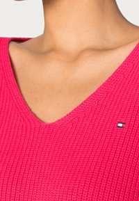 Gebreide roze V-hals trui met getextureerde stof en een klein geborduurd logo, met een geribd patroon en een subtiele glans.