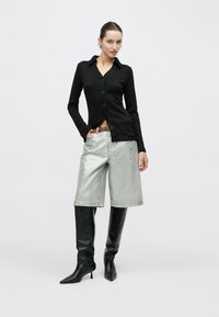 Μαύρο ριμπ μακρυμάνικο πουκάμισο με κουμπιά, ασημί μεταλλιζέ σορτς μέχρι το γόνατο και μαύρες μυτερές μπότες μέχρι το γόνατο. Ουδέτερο φόντο.