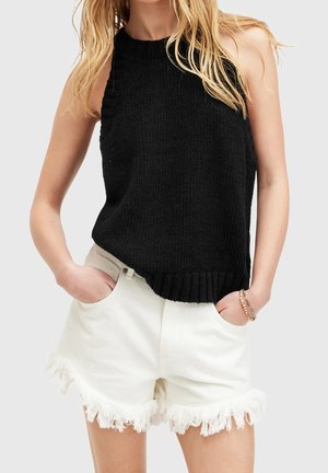 Blonde vrouw met een mouwloos zwart gebreid topje en witte denim shorts met rafels, met handen in de zakken en armbanden om de pols.