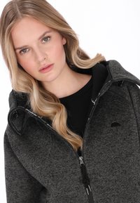 Sötétszürke cipzáros kapucnis pulóver texturált mintával, kettős fekete cipzárakkal és egy logóval a mellkason. Fekete belső bélés és kapucni.