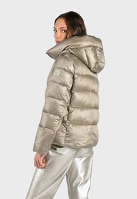 Lys beige pufferjakke med hætte, der har et quiltet design, skinnende overflade og et emblem på venstre skulder.