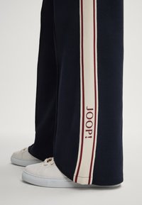 Marineblauwe sweatpants met een brede witte streep met een bordeauxrode accent, voorzien van de tekst "JOOP!" in bordeauxrood. Witte sneakers met ronde neuzen.
