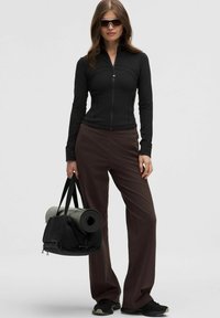 Schwarze Sportjacke mit Reißverschluss, braune weite Hose, schwarze Sportschuhe; hält eine schwarze Sporttasche und eine gerollte Yoga-Matte.