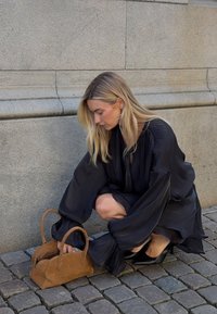 Melnā zīda blūze ar apjomīgām piedurknēm, apvienota ar brūnu zamša rokassomu. Iestatījums ir ar akmeņu bruģi un akmens sienu.