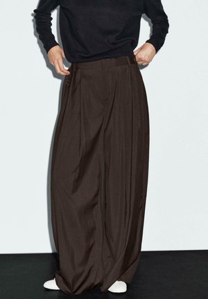 Pantalons marron à jambes larges en tissu doux, avec de profonds plis et une taille haute, associés à des chaussures blanches.
