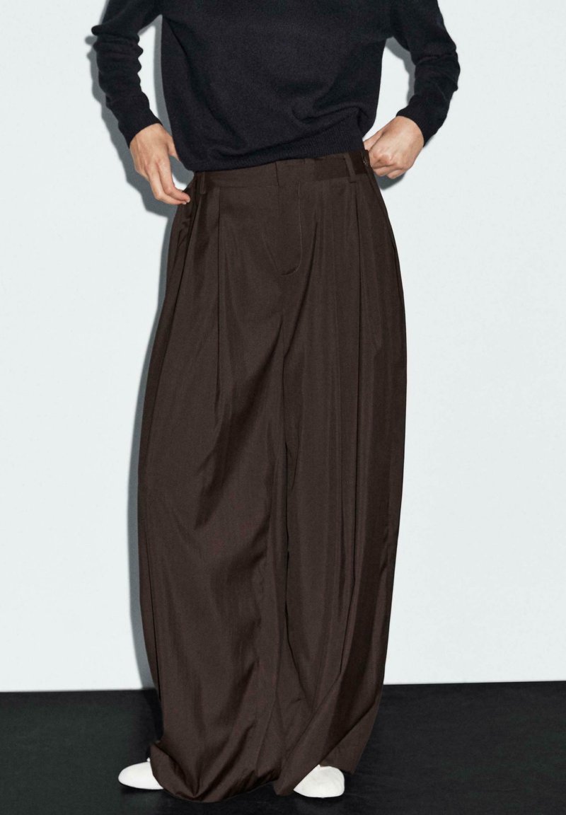 Pantalons marron à jambes larges en tissu doux, avec de profonds plis et une taille haute, associés à des chaussures blanches.