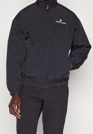 Mand iført en sort Sergio Tacchini lynlåstrøje og matchende sorte bukser med den ene hånd i lommen og synligt logo på brystet.
