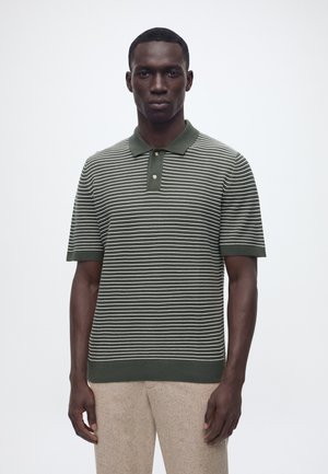 Mann trägt ein dunkelgrün-weiß gestreiftes Polohemd mit kurzen Ärmeln und beige Hose, steht vor einem einfarbigen hellen Hintergrund.