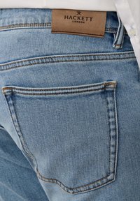 Svijetloplave traperice s natrag džepom u kojem je narančasti šav i smeđi kožni logotip Hackett London iznad struka.