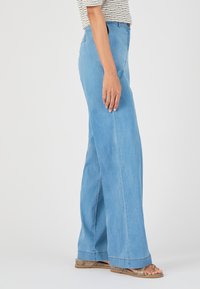 Pantalones de mezclilla de pierna ancha en un azul claro, con cintura alta, frente sin costuras, bolsillos laterales y dobladillo cosido. Combinados con sandalias planas y estampadas.