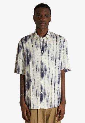 Giovane uomo che indossa una camicia tie-dye a maniche corte con bottoni, nera e bianca, abbinata a pantaloni beige, in piedi contro uno sfondo semplice.