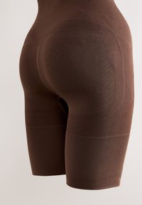 Približna snimka smeđih bešavnih kratkih hlača za oblikovanje koje ističu kontur šavove i teksturu tkanine na bokovima i bedrima.