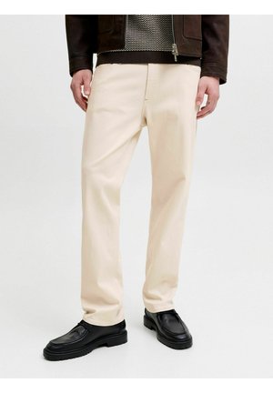 Homme portant un pantalon crème à coupe droite, des chaussures en cuir noires, une veste sombre et un pull à motifs, debout sur un fond blanc.