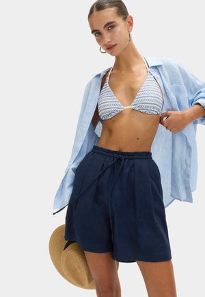 Młoda kobieta w niebieskim bikini z wzorem, luźnej jasnoniebieskiej koszuli i granatowych szortach, trzyma słomkowy kapelusz na jednolitym tle.