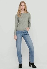Grauer Strickpullover mit Rundhalsausschnitt, kombiniert mit hellblauen Jeans. Das Modell trägt schwarze Absatzstiefeletten.
