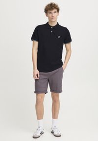Sort polo-shirt med krave, korte ærmer og et logo på venstre bryst. Sammen med opsmøgede grå shorts og hvide sneakers.